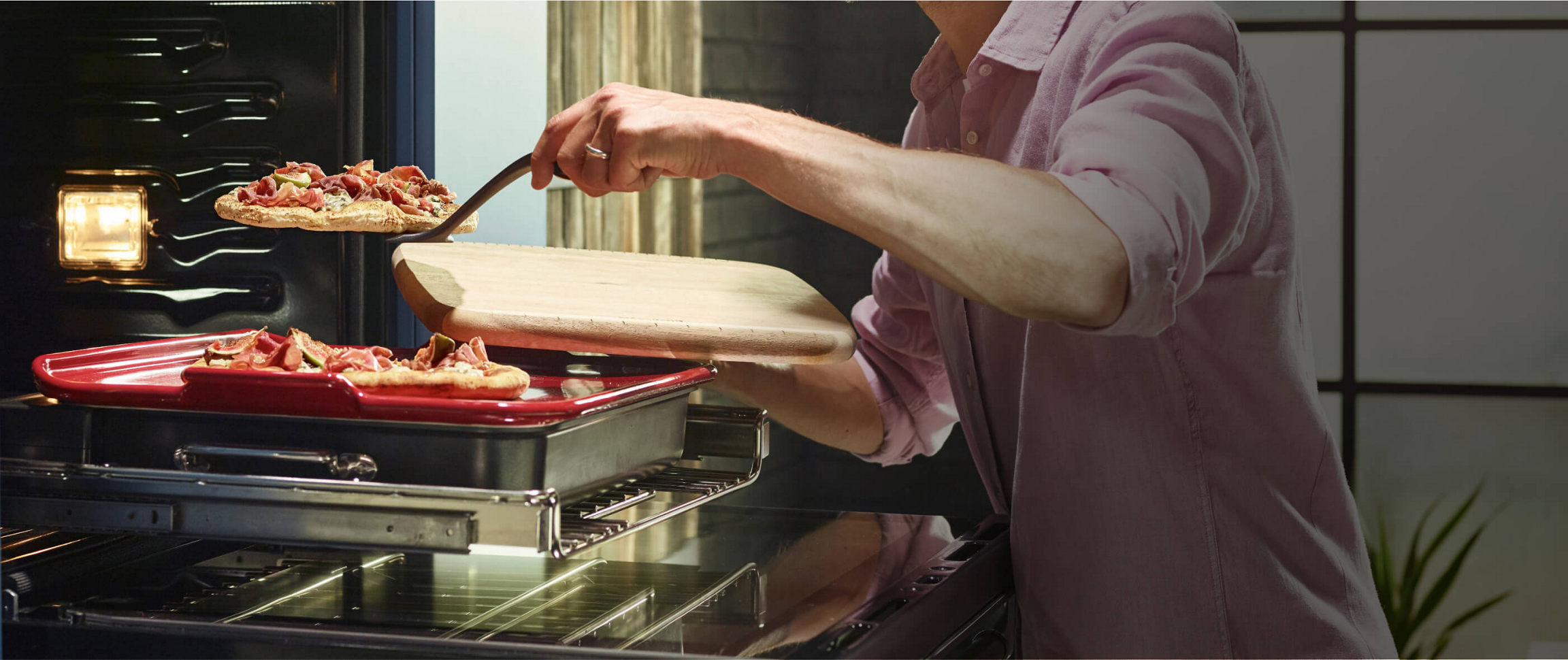 A Smart Oven Plus, open with the pizza stone attachment slid forward. A Maker transfers one of two gourmet pizza onto a cutting board for service.